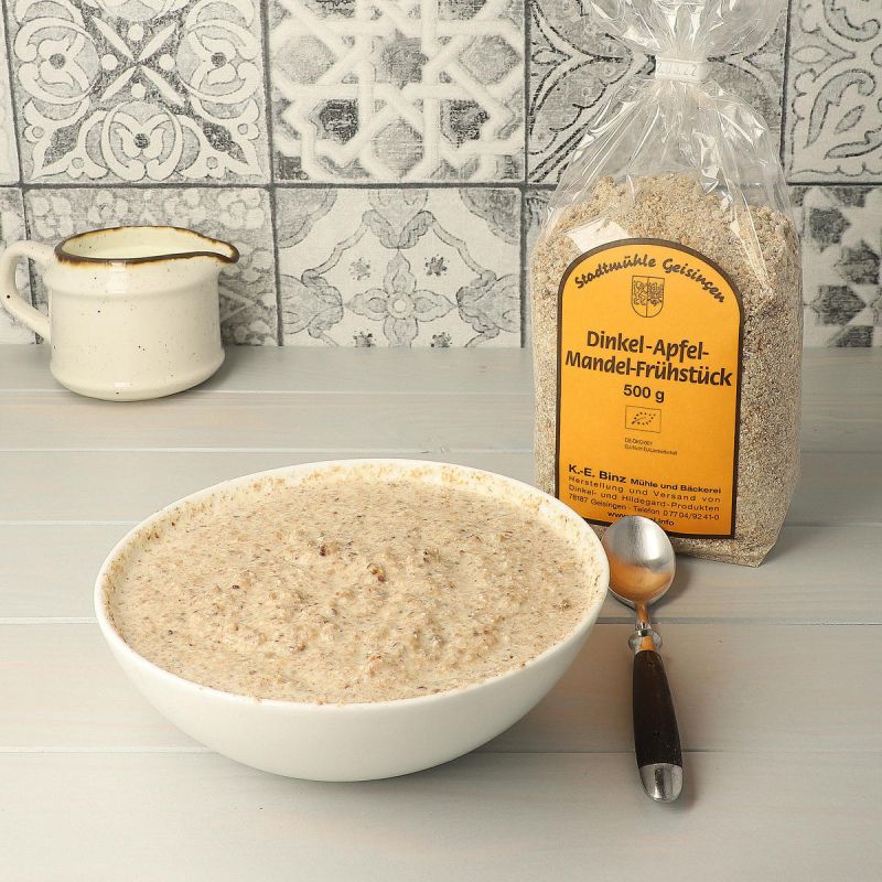 White bowl of pale grain porridge with a spoon, a small jug and a packet of spelt-apple-almond breakfast mix in front of patterned tiles