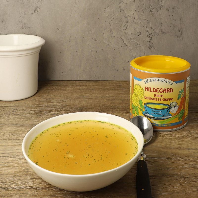 Light bowl of clear golden broth with herbs, a spoon beside it and a yellow tin of Hildegard clear delicacy soup