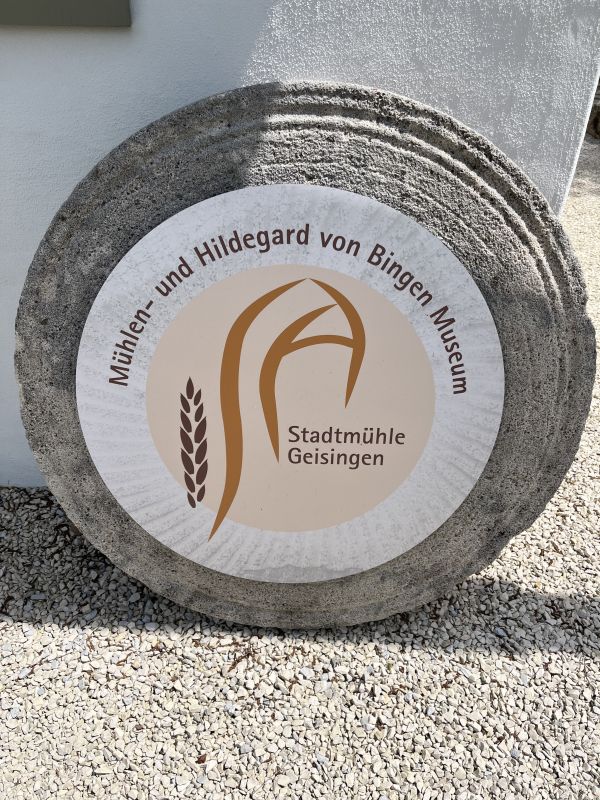 An old millstone bearing the inscription &ldquo;M&uuml;hlen- und Hildegard von Bingen Museum &middot; Stadtm&uuml;hle Geisingen&rdquo;, leaning against a white wall