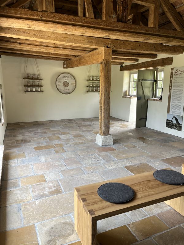 The exhibition room: stone floor, wooden pillar and beamed ceiling, shelves with jars and a round wall panel, a wooden bench with felt cushions in front