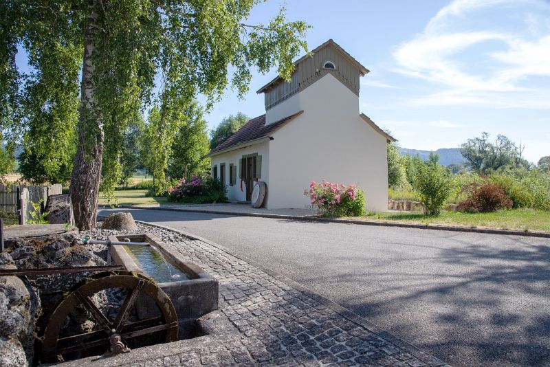 The white museum building with its timber upper storey seen from the road, a stone fountain trough with a small waterwheel in front, beside it a birch tree and roses in bloom